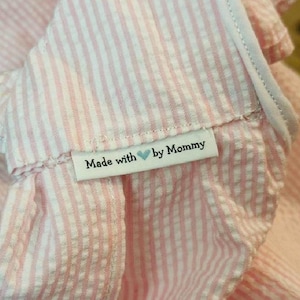 May include: A light pink and white seersucker fabric garment featuring a white rectangular clothing label sewn onto the seam. The label displays the black text Made with followed by a small blue heart icon and the word by Mommy.