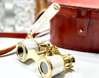 May include: A vintage gold-toned opera glass with white mother-of-pearl accents. The binoculars are next to a brown leather case with a red interior. A brown leather strap and two clear glass containers are in the background.
