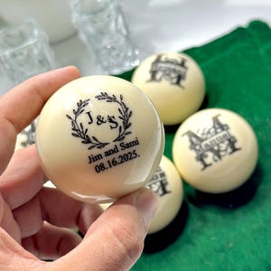 May include: Cream-coloured billiard balls with black custom text and graphics. One ball has the names "Jim and Sami" and the date "08.16.2025" inside a laurel wreath. Other balls display custom initials and designs. The balls are on a green felt surface.