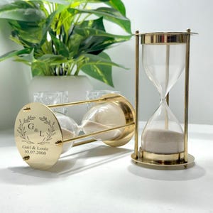 May include: Two gold-colored hourglasses with clear glass chambers filled with sand. One hourglass is upright, the other is lying on its side. The base of the hourglass has the engraved names "Gail & Louie" and the date "10.07.2000".