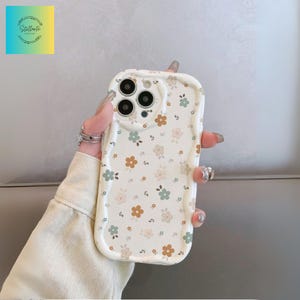 May include: A cream-coloured phone case adorned with a floral design of small brown, green, and pink flowers. The case has a wavy edge and a camera aperture. A logo with the text "Stellante" is in the upper left corner.