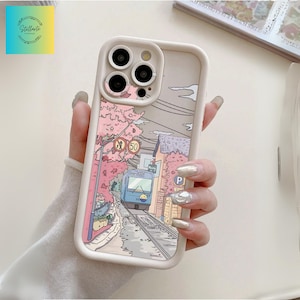 以下が含まれることがあります： ベージュ色の携帯電話ケース。桜の木のある街を走る電車のカラフルなイラストが描かれています。ケースには白い縁取りとカメラ用の切り抜きがあります。