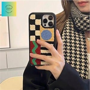 Op de afbeelding: Zwarte telefoonhoes met een kleurrijk, getextureerd ontwerp. De hoes heeft een dambordpatroon, strepen en geometrische vormen in rood, groen, blauw en beige. De hoes heeft een zwarte rand en een camera-uitsparing.