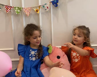 Puede incluir: Dos niñas pequeñas con vestidos azules y naranjas con bordados florales. El vestido azul tiene mangas con volantes. Un peluche rosa y globos están en primer plano. Una colorida pancarta cuelga en el fondo.