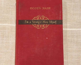 May include: A vintage red hardcover book titled "I'm a Stranger Here Myself" by Ogden Nash. The title is printed in gold lettering on a black band across the cover. The book is on a neutral-colored, textured surface.