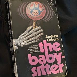 May include: Vintage paperback book titled "The Babysitter" by Andrew Coburn. The cover features a black background with a pink and white baby rattle held by a skeleton hand. The title is in pink.