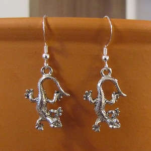 May include: A pair of silver-colored lizard earrings. Each earring features a detailed lizard charm with textured skin and outstretched limbs, suspended from a simple hook. The earrings are designed for pierced ears and are displayed against a warm, neutral background.