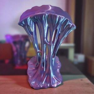 Op de afbeelding: Een abstracte sculptuur met vloeiende, organische vormen. Het stuk heeft een afgeronde basis die overgaat in verticale, open structuren, eindigend in een afgeronde top. De kleur verschuift tussen paars en blauw, waardoor een iriserend effect ontstaat.