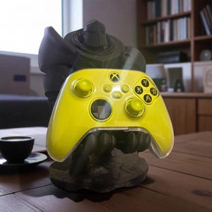 May include: A bright yellow Xbox controller rests in a dark gray, stylized holder shaped like a figure. The controller has black buttons and a clear plastic shell. The holder sits on a wooden surface.