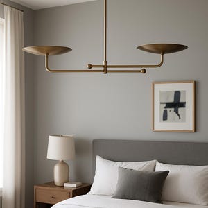 Op de afbeelding: Een moderne messing kroonluchter met twee cirkelvormige kappen, opgehangen aan het plafond. De armatuur bevindt zich boven een bed met wit beddengoed, een grijs hoofdbord en een ingelijst abstract kunstwerk. Een bedlampje is ook zichtbaar.