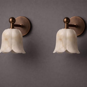 Modern brass wall sconce set with alabaster stone shade, minimalist bedroom wall lighting on grey background Mid century brass wall lights with white alabaster tulip shades, luxury wall sconce pair for bedroom decor Set of two brass wall sconces with natural alabaster shades, elegant modern wall lighting fixture Minimal brass wall lamps with alabaster shade, bedside wall lighting pair on neutral grey wall Luxury brass wall sconce pair with alabaster tulip glass shade, modern wall lighting.