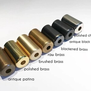 May include: A collection of cylindrical metal objects in various finishes, including antique patina, polished brass, brushed brass, raw brass, blackened brass, antique black, and polished chrome. Each cylinder has a hole at both ends, and the names of the finishes are labeled.