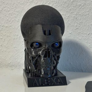 Op de afbeelding: Een zwarte schedelvormige luidsprekerstandaard met gloeiende blauwe ogen. De standaard is gemaakt van een donker materiaal en heeft het woord "ALEXA" op de basis. De luidspreker zelf is bedekt met een zwarte mesh-stof.