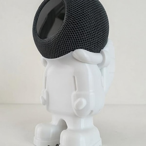Könnte beinhalten: Eine weiße Figur in Form eines Astronauten, der einen schwarzen Lautsprecher hält. Der Lautsprecher hat ein Mesh-Design und ein dunkles Inneres. Die Astronautenfigur hat ein cartoonartiges Design mit großen Füßen und Händen.
