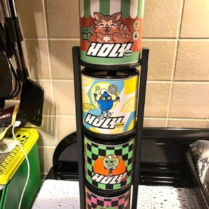 Puede incluir: Un soporte de metal negro que sostiene cuatro recipientes cilíndricos de colores. Cada recipiente presenta un personaje de dibujos animados diferente y la palabra "HOLY". El recipiente superior tiene un gato, el segundo una pelota de tenis, el tercero una manzana y el cuarto un limón.