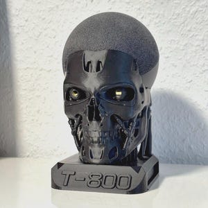 Op de afbeelding: Zwarte schedelvormige luidsprekerstandaard met de tekst "T-800" op de basis. De luidspreker heeft een grijze stoffen bovenkant en felgele ogen. De schedel is een gedetailleerde replica van een Terminator.