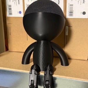 Könnte beinhalten: Eine schwarze, 3D-gedruckte Figur mit einem abgerundeten Kopf, der mit dunkelgrauem Stoff bedeckt ist. Die Figur hat einen kugelförmigen Körper, kleine Arme und gegliederte Beine, die auf einem Metallregal sitzen. Der Hintergrund umfasst Kartons mit Produktinformationen.