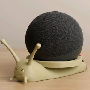 May include: A novelty speaker stand in the shape of a snail, coloured light green. It supports a dark grey, spherical speaker. The snail has a smiling face and two antennae. The speaker is a smart home device.