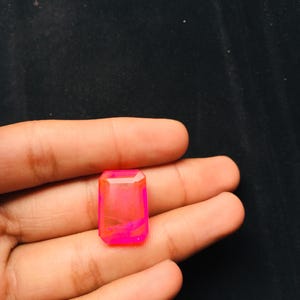 May include: A rectangular, emerald-cut gemstone featuring a vibrant, translucent neon pink hue with subtle internal color variations. The stone has faceted edges that catch the light, creating a bright, glowing appearance against the dark background.