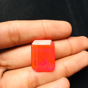 May include: A rectangular, faceted gemstone with a vibrant neon orange and pink hue. The stone features a smooth, polished surface with internal dark, branch-like inclusions visible throughout the translucent, glowing orange body of the gem.