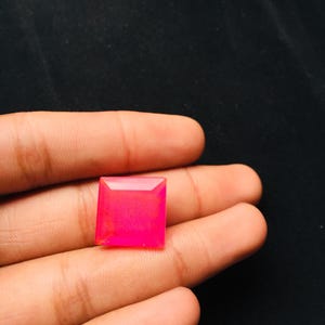 May include: A square-cut, vibrant hot pink gemstone with a faceted surface that reflects light. The stone features sharp, clean edges and a polished finish, displaying a consistent, intense magenta hue throughout its entire square structure.