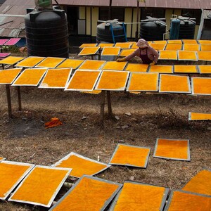 Puede incluir: Un grupo de personas trabajando en una ladera, secando hojas de papel naranja en marcos de madera. El papel est&aacute; dispuesto en filas en los marcos, y las personas trabajan para asegurar que el papel se seque uniformemente.