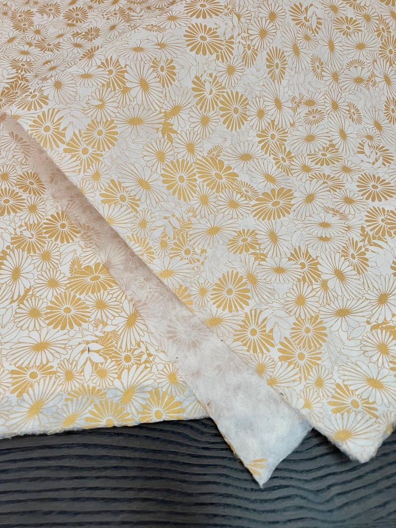 K&ouml;nnte beinhalten: Wei&szlig;es Seidenpapier mit einem goldenen Blumenmuster. Das Muster zeigt kleine, stilisierte G&auml;nsebl&uuml;mchen.