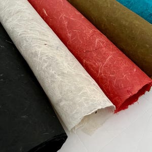 May include: A collection of four rolled sheets of handmade paper featuring a fibrous, textured surface. The colors include black, off-white, red, olive green, and bright blue, each displaying visible natural plant fibers throughout the material.