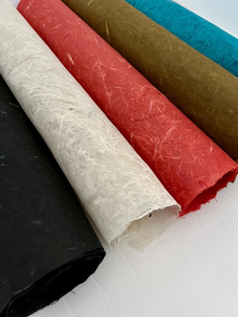 May include: A collection of four rolled sheets of handmade paper featuring a fibrous, textured surface. The colors include black, off-white, red, olive green, and bright blue, each displaying visible natural plant fibers throughout the material.