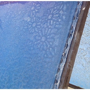 May include: Close-up of a blue fabric with a floral pattern. The fabric is stretched over a wooden frame and secured with staples. The floral design is subtly embossed on the fabric's surface. The image is well-lit, highlighting the texture and details.