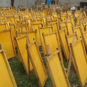 May include: A large collection of rectangular wooden frames, each containing a bright yellow sheet of material, arranged in rows and supported by bamboo poles. The frames are positioned upright to allow the yellow sheets to dry in the open air.
