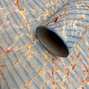 May include: A rolled-up sheet of paper with a marbled design. The paper has a light blue background with a ribbed texture. The marbling features gold, red, and white veins, creating a decorative pattern. The rolled edge reveals the paper's interior.