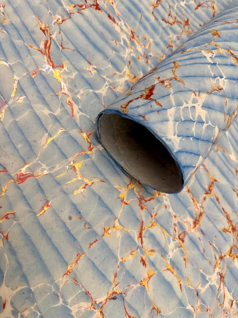 May include: A rolled-up sheet of paper with a marbled design. The paper has a light blue background with a ribbed texture. The marbling features gold, red, and white veins, creating a decorative pattern. The rolled edge reveals the paper's interior.