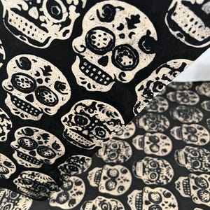 May include: A black fabric sheet featuring a repeating pattern of off-white sugar skulls. The skulls are decorated with intricate floral and geometric details, including circular eye sockets and teeth, printed in a high-contrast black and white style.