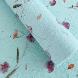 Könnte beinhalten: Eine Rolle hellblaues handgeschöpftes Papier mit einer strukturierten Oberfläche, in die verstreute dunkelrosa und orangefarbene getrocknete Blätter sowie dünne braune Stängel eingebettet sind. Das Papier wirkt faserig und organisch.