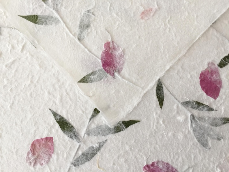 May include: Close-up of handmade paper with embedded pink flower petals and green leaves. The paper has a textured, organic appearance, with visible fibers and a slightly off-white color. The design suggests a natural, botanical theme.