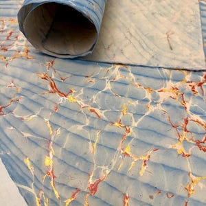 May include: A close-up of marbled paper in shades of blue, white, gold, and red. The paper is partially rolled, revealing a textured surface with a wave-like pattern. The gold and red veins create a striking contrast against the blue.