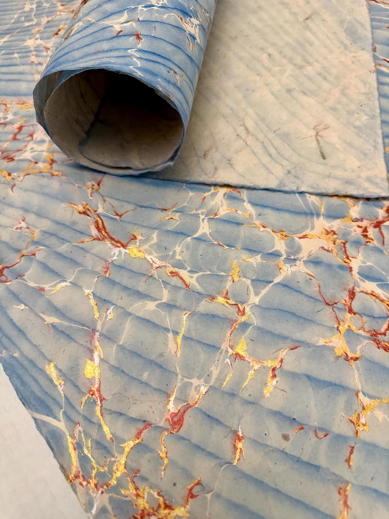 May include: A close-up of marbled paper in shades of blue, white, gold, and red. The paper is partially rolled, revealing a textured surface with a wave-like pattern. The gold and red veins create a striking contrast against the blue.