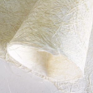 May include: A sheet of off-white, translucent handmade paper featuring a visible, fibrous, and tangled texture throughout its surface. The paper is partially rolled, showcasing its delicate, uneven edges and a light, cream-colored appearance.