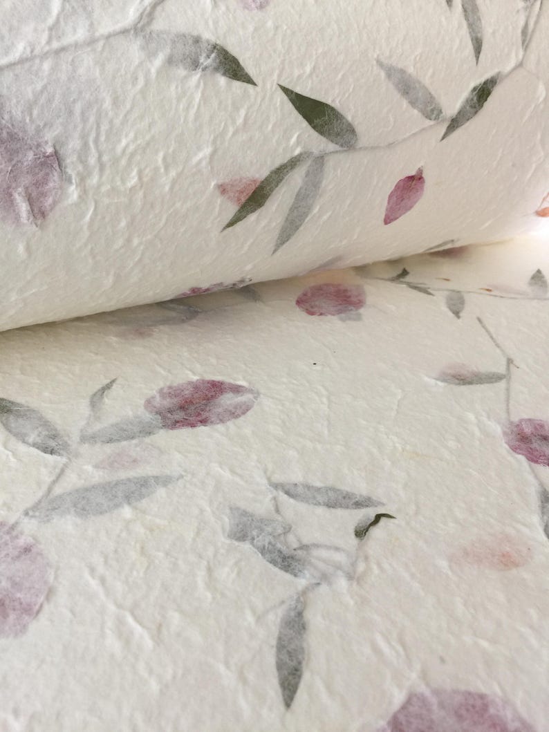 May include: Close-up of handmade paper with a floral design. The paper is white with a textured surface. The floral pattern features purple flowers and gray-green leaves. The paper is partially rolled, revealing the pattern.