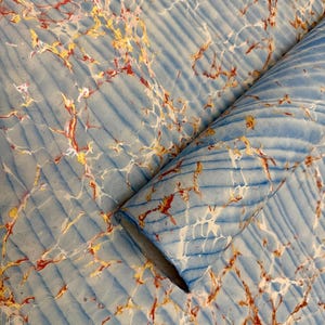 May include: A roll of marbled paper with a light blue background and diagonal ribbed texture. The paper features a pattern of gold, red, and white veins, creating a unique and artistic design. The paper is rolled up, showing the inside.