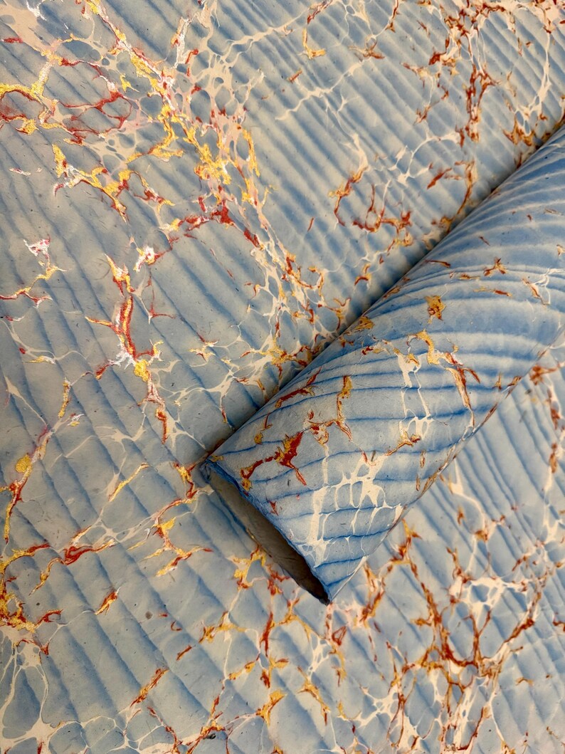 May include: A roll of marbled paper with a light blue background and diagonal ribbed texture. The paper features a pattern of gold, red, and white veins, creating a unique and artistic design. The paper is rolled up, showing the inside.
