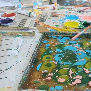 May include: A craft table with paint supplies. A tray of swirling blue, green, and pink paint is in the foreground. Paint tubes, paint palettes with various colors, and paintbrushes are scattered on the table, suggesting a painting activity.