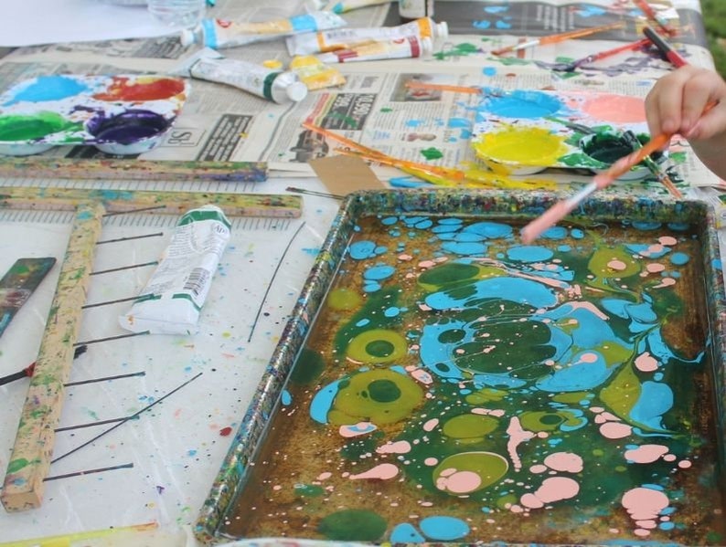 May include: A craft table with paint supplies. A tray of swirling blue, green, and pink paint is in the foreground. Paint tubes, paint palettes with various colors, and paintbrushes are scattered on the table, suggesting a painting activity.