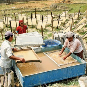 May include: People working outdoors, crafting paper. Workers are seen in a field with large blue tubs filled with water. They are using wooden frames to create paper. In the background, sheets of paper are drying on wooden frames.