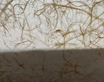 May include: A sheet of translucent white paper featuring an embedded pattern of thin, light brown fibrous strands that create a tangled, organic web across the surface, with a distinct horizontal shadow line dividing the upper and lower sections.