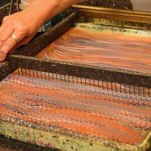 May include: A rectangular marbling tray containing a liquid surface with a swirling orange, blue, and white pattern. A metal comb with thin vertical tines is positioned across the tray, creating a repeating wave design in the paint.
