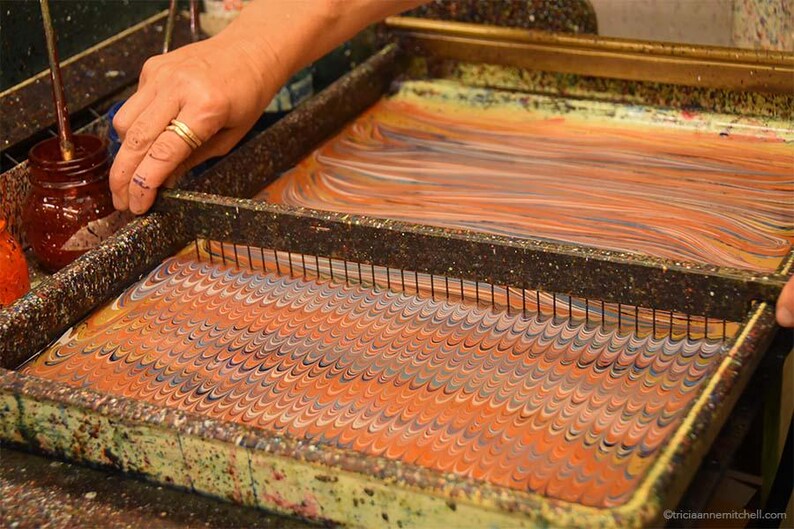 May include: A rectangular marbling tray containing a liquid surface with a swirling orange, blue, and white pattern. A metal comb with thin vertical tines is positioned across the tray, creating a repeating wave design in the paint.
