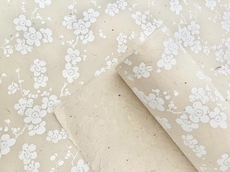 Puede incluir: Un rollo de papel de regalo beige con un patr&oacute;n floral blanco. El papel est&aacute; hecho de materiales reciclados y tiene una superficie texturizada.
