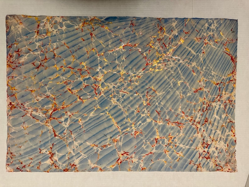 May include: Rectangular sheet with a marbled design. The background is a light blue with diagonal lines. The marbled pattern features red, yellow, and white veins, creating a unique and textured appearance. The edges are slightly uneven.